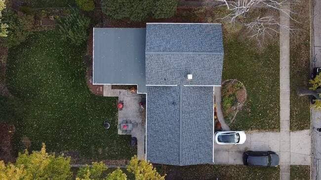 Aerial top-down view of a suburban L-shaped house featuring a newly installed dark gray asphalt shingle roof on the main section and a lighter gray flat roof on an extension. The image shows the entire property, including the green lawn, a patio area, and two cars parked in the driveway.